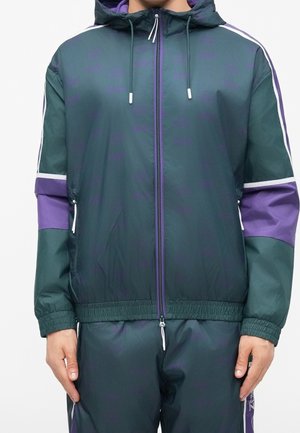 Osoba nosząca ciemnozieloną i fioletową bluzę dresową z kapturem, z zamkiem z przodu i białymi sznurkami, ręce spoczywają po bokach.