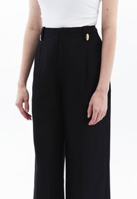 Hoge zwarte broek met plooien, met gouden knopen bij de taille en een wijde pijp-silhouet. Gladde stoftextuur.
