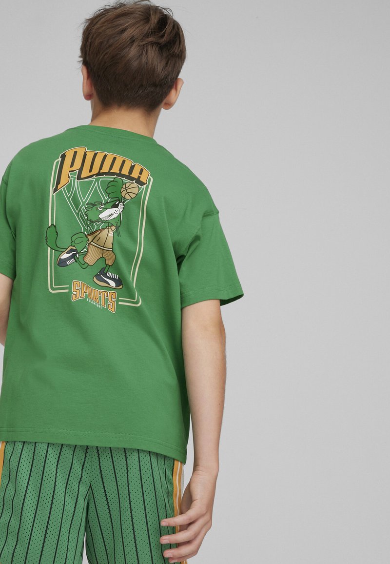 Maglietta in cotone verde con un personaggio dei cartoni animati che gioca a basket sul retro, con il testo "Puma" e "Sports". Fit casual e maniche corte.