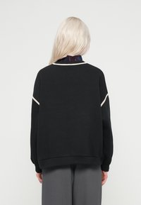 Μαύρη μπλούζα με μακριά μανίκια, με λείο ύφασμα και κρεμ ραφές κατά μήκος των ώμων και του λαιμού.