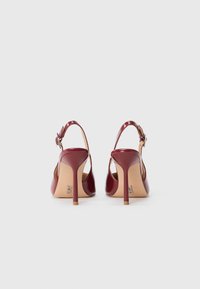 Tacchi alti slingback in pelle verniciata bordeaux con sottili tacchi a stiletto, cinturini alla caviglia regolabili e suole beige lisce, mostrati da dietro.