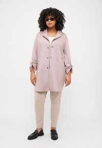 Light pink suede trench coat with a collar, double-breasted button closure, and rolled sleeves paired with beige trousers and black loafers.