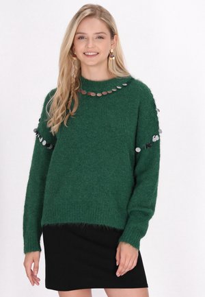 Ung kvinde med langt blondt hår iført grøn fluffy sweater med sølv palietter og sort nederdel, smilende mod en hvid baggrund.