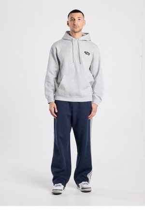 Grauer Hoodie mit einer Fronttasche und Kordelzügen, kombiniert mit marineblauen Hosen mit weißen Seitenstreifen. Das Modell steht aufrecht.