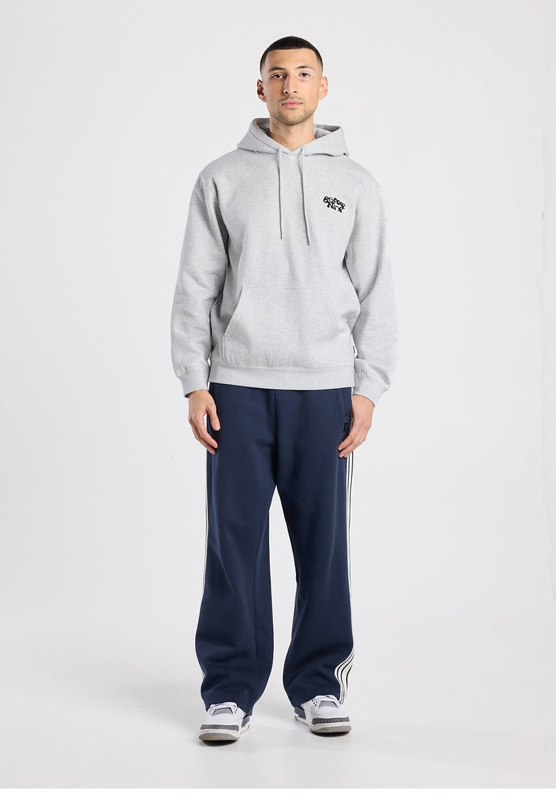 Sweat à capuche gris avec une poche avant et des cordons de serrage, associé à un pantalon bleu marine avec des bandes blanches sur les côtés. Le mannequin se tient droit.