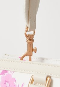 Clip metallico rotondo dorato attaccato a una maniglia chiara, fissato a una borsa bianca con motivi floreali rosa e cuciture a contrasto.