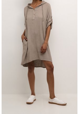 Femme portant une robe beige ample à capuche avec manches retroussées et baskets blanches, debout devant un fond clair uni