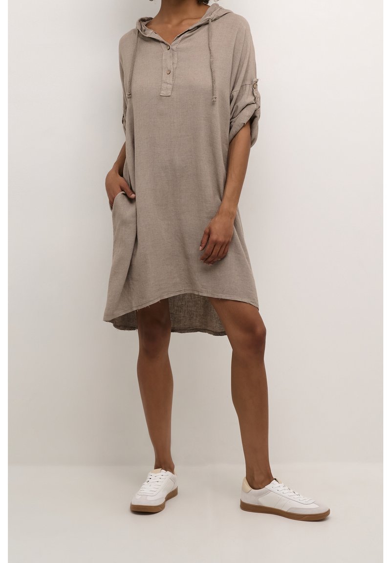 Femme portant une robe beige ample à capuche avec manches retroussées et baskets blanches, debout devant un fond clair uni