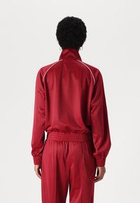 Red satin track jacket with white piping, high collar, and elastic cuffs. Relaxed fit, paired with coordinating pants featuring an elastic waistband.