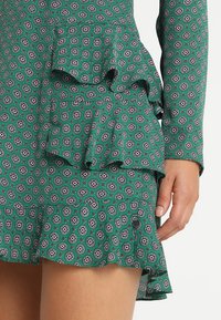 Vihreä mekko, jossa on kuvioitu design, röyhelökerrokset, pitkät hihat ja teksturoitu kangas. Huomionarvoiset yksityiskohdat sisältävät geometrisia muotoja ja yksityiskohtia.