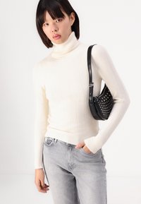 Un pull col roulé à côtes crème associé à un jean gris clair. Un petit sac à main noir avec des clous est porté sur le corps.