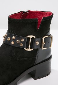 Black suede ankle boot with a gold-studded leather strap and buckle. Features a red leather interior and a chunky low heel.