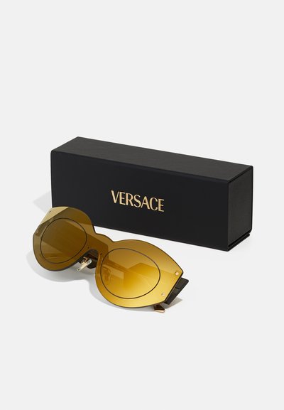 Versace päikeseprillid suurte kollaste läätsedega, geomeetrilise kujuga ja kuldsete detailidega, asetatud musta bränditud kasti kõrvale.
