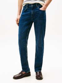 Jeans de ganga em azul escuro com um corte reto, apresentando cinco bolsos, passadores de cinto e detalhes de costura discretos, combinados com sapatos mocassim castanhos.
