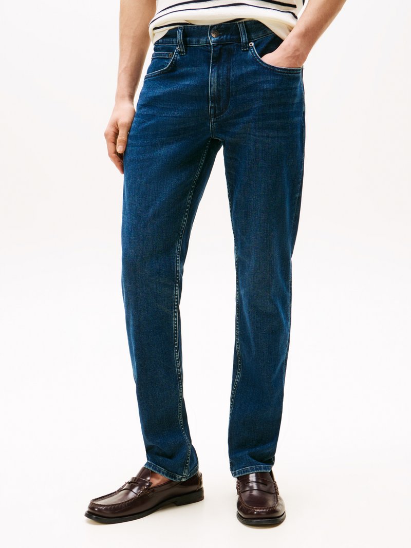Jeans de ganga em azul escuro com um corte reto, apresentando cinco bolsos, passadores de cinto e detalhes de costura discretos, combinados com sapatos mocassim castanhos.