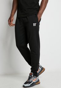 Mallas negras con el logo "11°" en blanco, cintura elástica y bolsillos laterales. Combinadas con zapatillas deportivas multicolores que tienen una suela de aire visible.