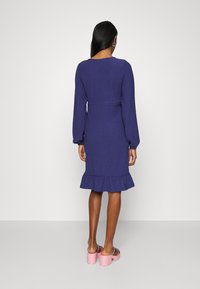 Fialové šaty s dlouhým rukávem, rozšířenými lemy a přiléhavým pasem. Hladká textura s jemným vzorem. V kombinaci s růžovými platformovými sandály.