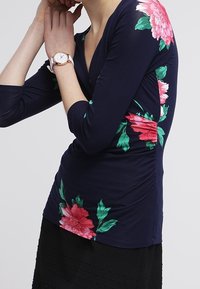 Mörkblå långärmad topp med rosa och gröna blommönster på ärmarna, design med v-ringning och draperade detaljer på sidan. Slät tygkänsla.