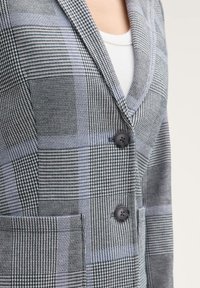 Šedý a modrý kostkovaný blazer vyrobený z texturované látky, s klopami, dvěma knoflíky a předními kapsami.
