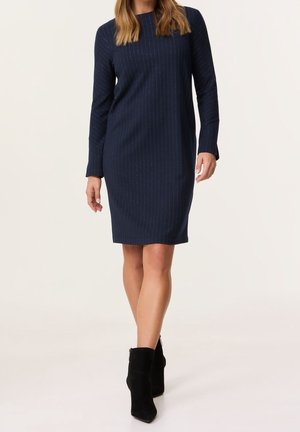 Kobieta ubrana w granatową, długą sukienkę do kolan z delikatnymi pionowymi paskami i czarne botki do kostki, stojąca na białym tle.