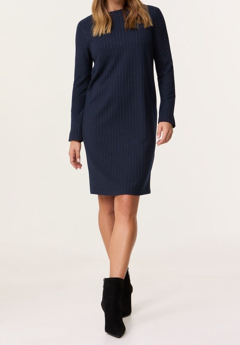 Femme portant une robe bleu marine à manches longues, longueur genou, avec de fines rayures verticales et des bottines noires, debout sur un fond blanc.