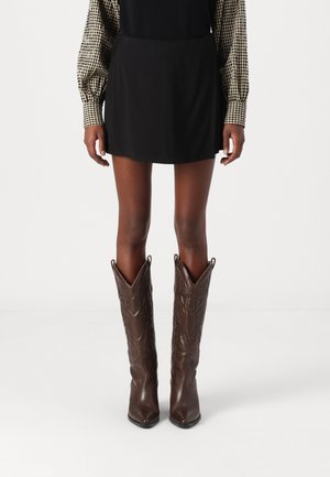 Brown leather cowboy boots with pointed toes and decorative stitching. Worn with a black skirt and patterned long-sleeve shirt.