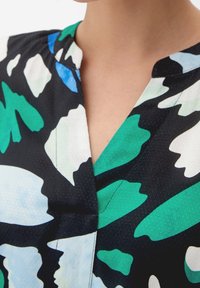 Lyhythihainen mekko, jossa on musta pohja, vihreitä, sinisiä ja valkoisia lehtikuvioita, sekä V-pääntie, jossa on minimaalista ompelutusta.