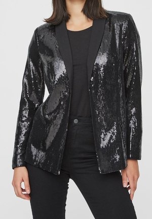 Woman wearing a black sequin blazer over a black top and black pants, standing with hands relaxed at sides.