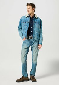 Giacca in denim azzurro con colletto in ecopelliccia crema, abbinata a una camicia blu navy e jeans azzurri. Stivali marroni con lacci completano l'outfit.