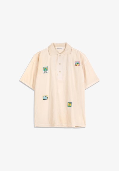 Polo beige clair à manches courtes avec quatre écussons carrés colorés présentant des motifs abstraits et de dessins animés sur le devant.