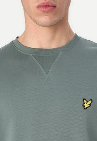 Felpe verdi realizzate in morbido tessuto, con collo a costine e un piccolo logo di un uccellino giallo sul petto. Design semplice e informale.