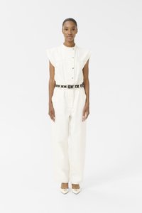 Combinaison en denim couleur crème avec des manches courtes et structurées, des boutons à l'avant et une ceinture noire avec des éléments en métal argenté. Le modèle porte des talons beiges à bout pointu.