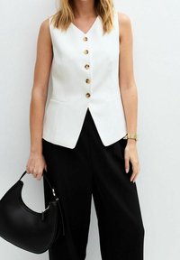 Bílý bezrukávový blazer s výstřihem do V, zlatými knoflíky a bočními kapsami, doplněný černými kalhotami s širokými nohavicemi a černou kabelkou.