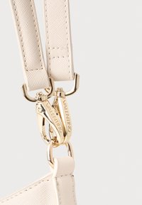 Svijetlo bež remen od teksturirane tkanine, s zlatnim metalnim kopčama. Metalne dijelove krasi izgravirani natpis "Valentino" za dodatni detalj.