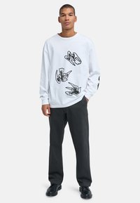 Jonge man die een wit shirt met lange mouwen draagt met zwarte vogelillustraties, zwarte broek en zwarte schoenen tegen een effen achtergrond.