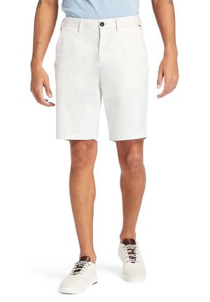 Homme portant un short blanc coupé au genou, une chemise bleu clair rentrée, et des baskets blanches avec des lacets brun foncé, debout, bras détendus.