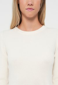 Ribbed long-sleeve top in off-white knit fabric, featuring a round neckline and subtle texture, designed for a fitted silhouette.