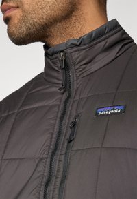 Μπουφάν πατιγιέ Patagonia σε σκούρο γκρι, με ψηλό γιακά, φερμουάρ μπροστά και διακριτικό σήμα με μπλε και κόκκινες λεπτομέρειες.