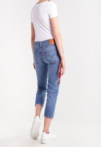 Ljusblå denimjeans med cropped passform, med röd blommig broderi på vänster ben. Matchas med en vit tajt t-shirt och vita sneakers.