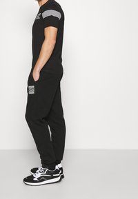 Svart t-shirt med vita randiga detaljer på ärmarna, parat med svarta joggers med en mönstrad ficka. Bärs med svartvita sneakers.