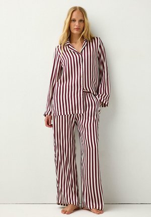 Jonge vrouw staat blootsvoets, draagt een bijpassend overhemd met lange mouwen en knopen en een wijde broek met verticale kastanjebruine en witte strepen.