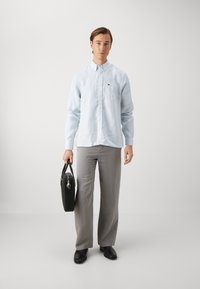 Hellsblau und weiß gestreifte Button-Up-Bluse mit einer Brusttasche, graue Anzughose und schwarze Lederschuhe, hält eine schwarze Aktentasche.