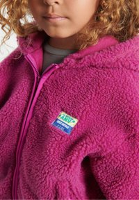 Fleecejakke i levende pink, med en struktureret overflade, fuld lynlås og en logo-patch mærket "SUPER VINTAGE" på brystet.