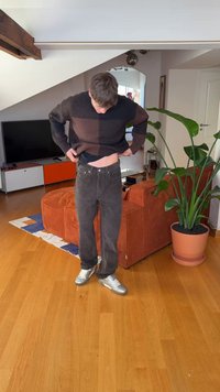 Brązowo-czarny sweter w paski, czarne jeansy i srebrne buty. Wnętrze z zieloną rośliną w doniczce i pomarańczową kanapą.