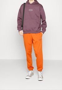 Lila hoodie med ficka fram och logotyp, matchad med orangea joggers som har en logotyp. Vita sneakers med zebramönster.