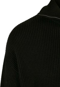 Sweat-shirt noir en tricot côtelé avec un col haut et une fermeture éclair sur toute la longueur du col. La texture est épaisse et tissée.