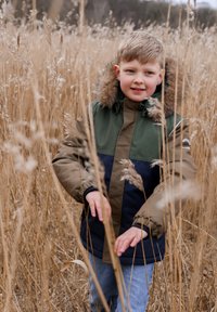 Prošívaná bunda v barvách olivově zelené, námořnické modré a béžové s kapucí lemovanou kožešinou; nošená přes modré džíny ve vysoké suché trávě.