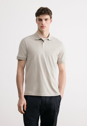 Jeune homme portant un polo beige et un pantalon noir, debout devant un fond clair uni avec une expression neutre.