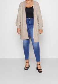 Cardigan lungo beige con vestibilità rilassata, apertura frontale e polsini a coste, abbinato a jeans skinny blu scuro e sandali neri con tacco largo.