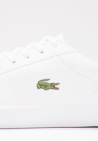 Baskets blanches avec des lacets blancs et un petit logo de crocodile vert brodé sur le côté.